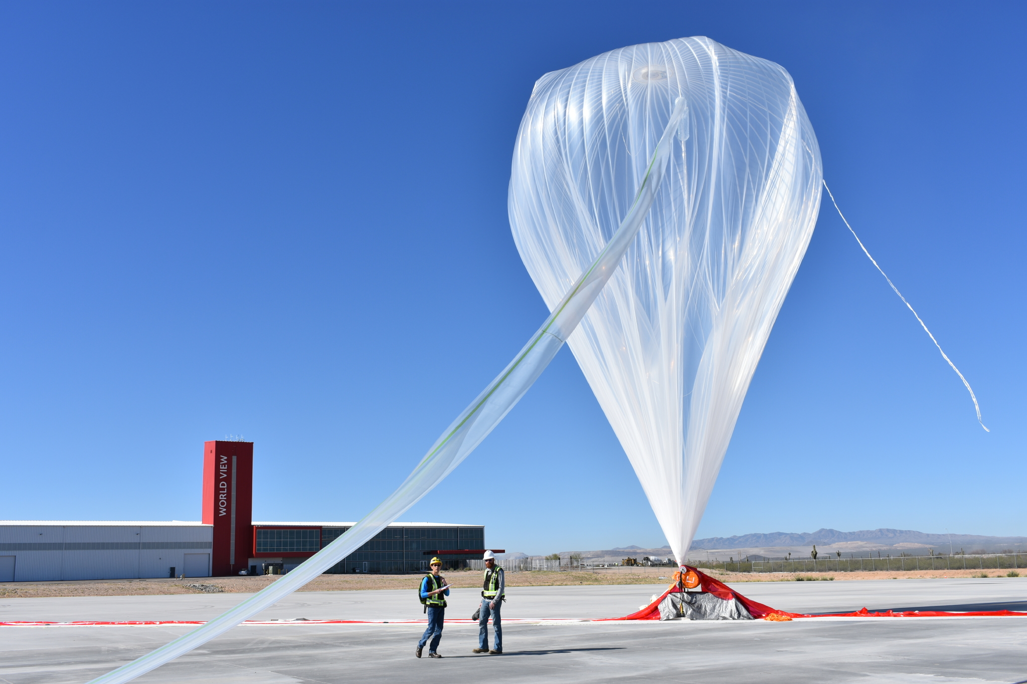two crew beside an inflated high-altitude balloon, with a building labeled World View in the background