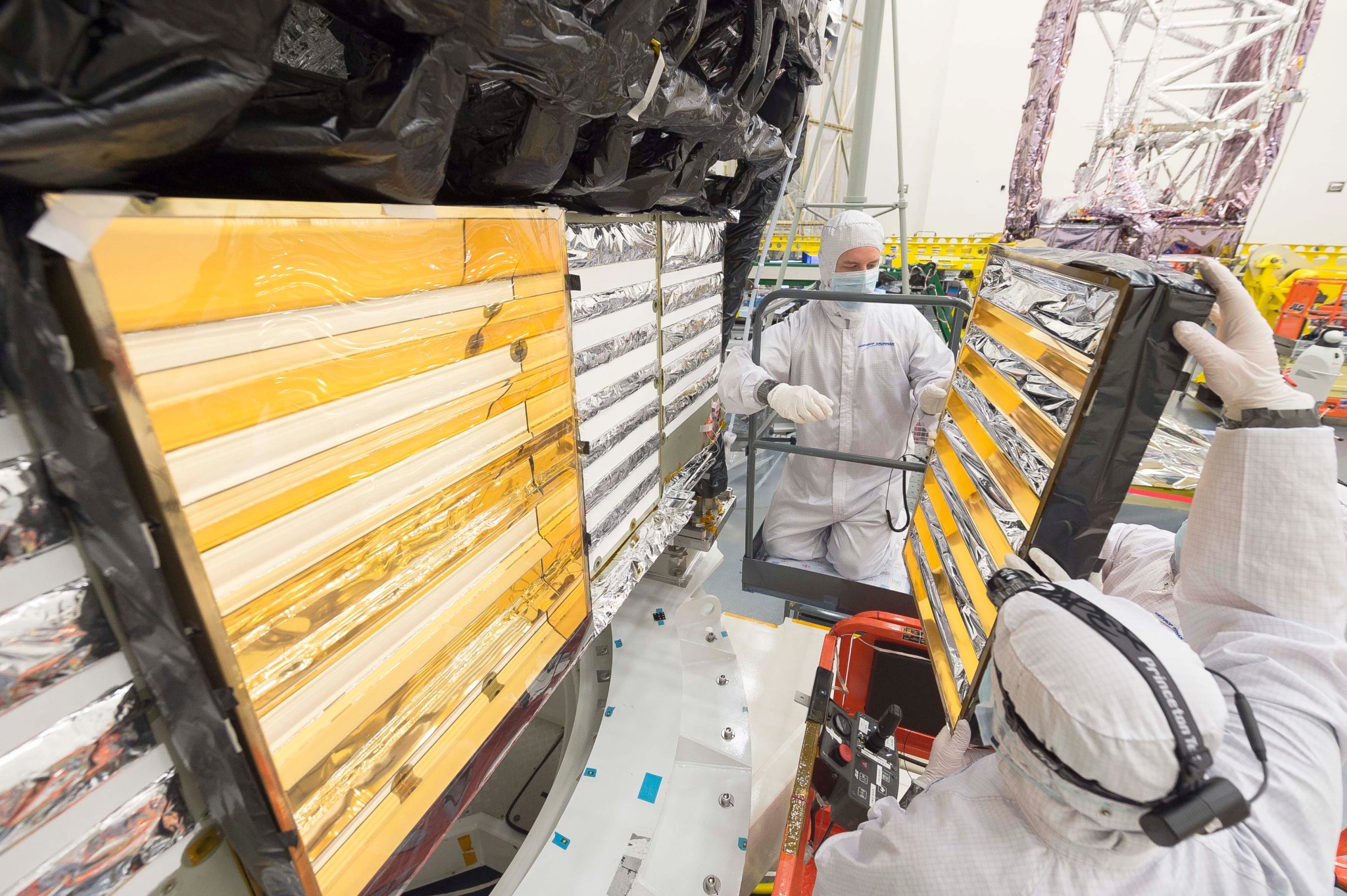 Engineers carefully hold onto a gold-plated baffle as they use a scissor lift to access the back of the ISIM.