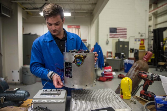 A student in a blue static jacket holds a piece of a rocket payload.