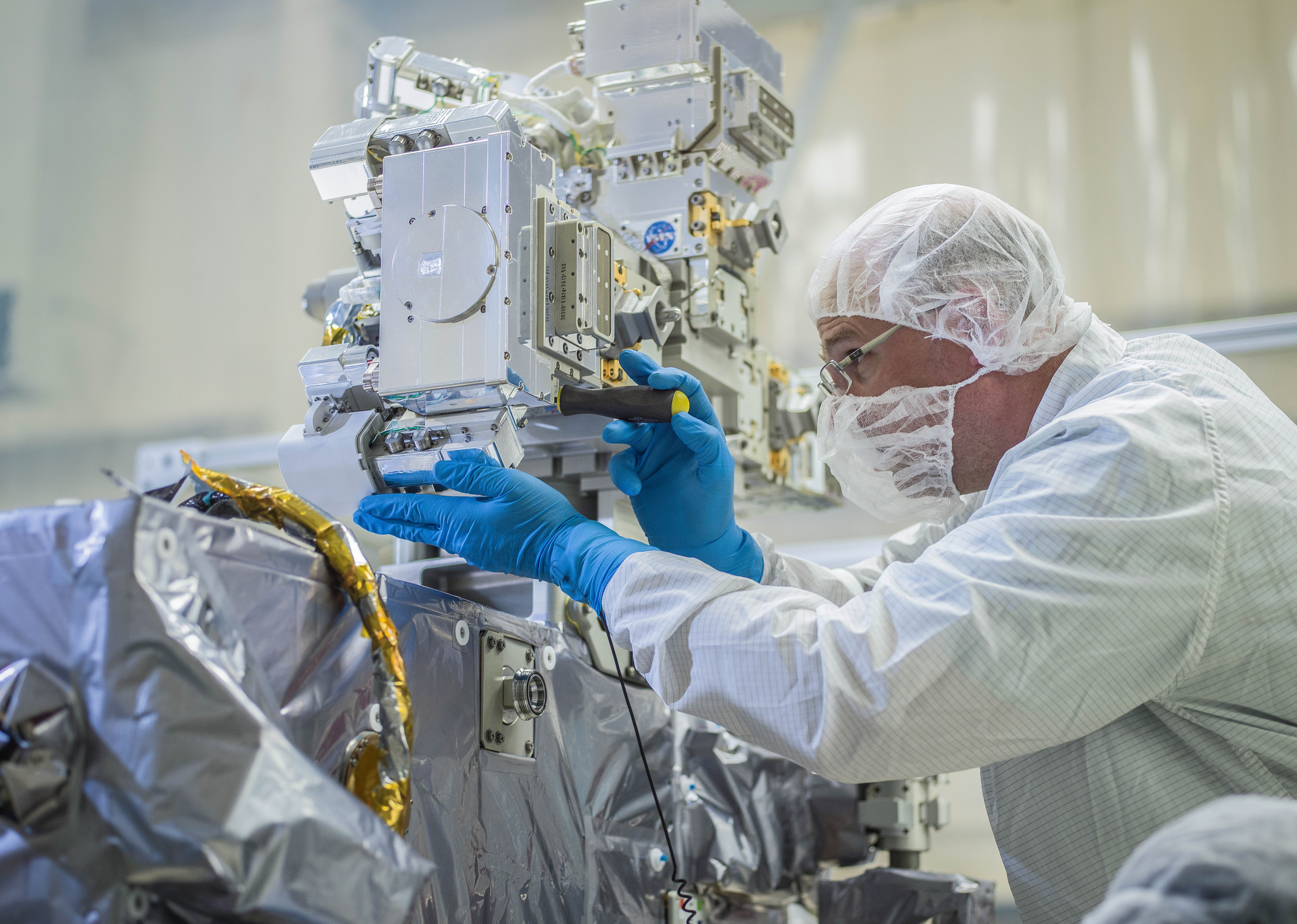 A man in coveralls, a white surgical mask, blue gloves, and safety glasses leans forward to work on a scientific instrument, making an adjustment with a screwdriver. He is in a clean room with white walls in the background.