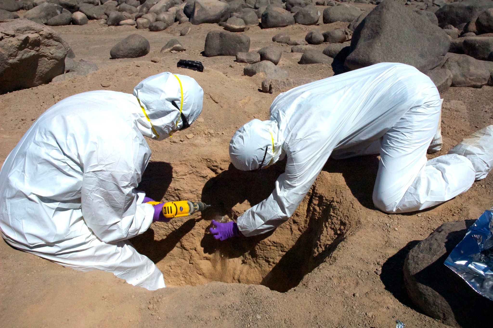 Two researchers collecting samples from a hole in the ground in the Atacama Desert using a drill.