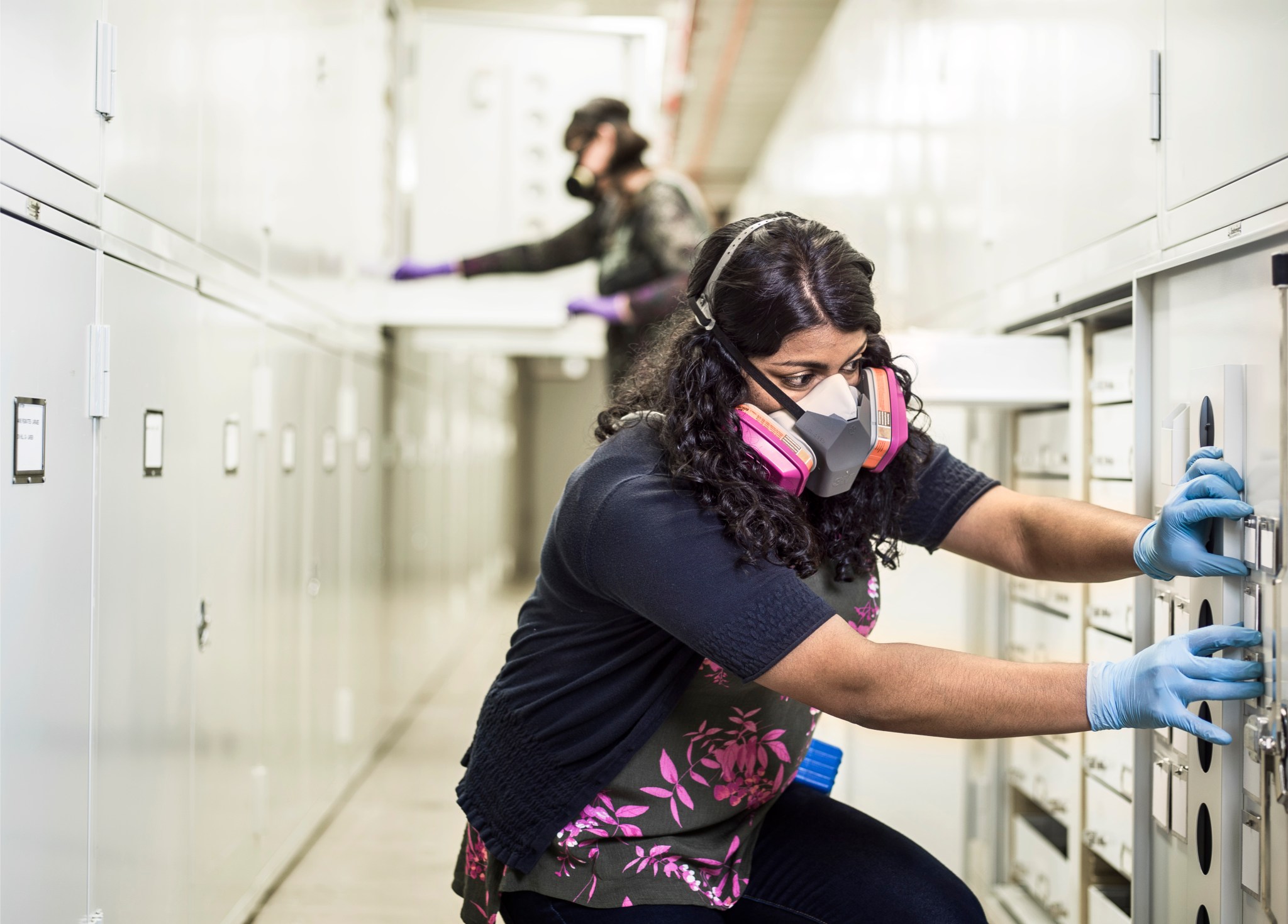 Woman with dark tan skin and curly black hair wears a pink gas mask and blue gloves. She is touching a specimen-storage cabinet. 