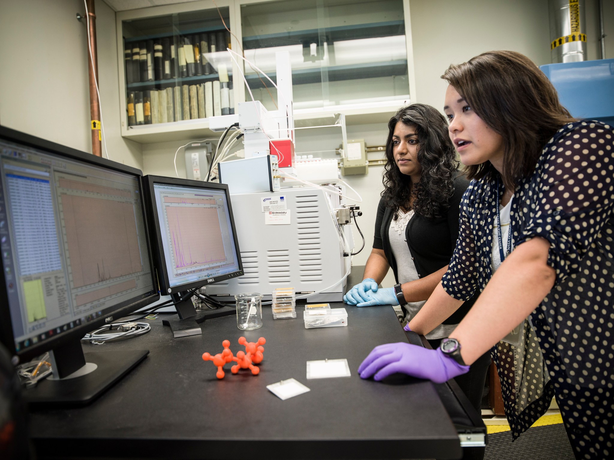 Two women, the forefront woman has tan skin, straight dark brown hair and wears a black and white polkadot shirt and purple gloves, the woman in the back has dark tan skin and curly black hair. She wears a black cardigan and blue gloves. The women are looking at two computer monitors displaying graphs. 