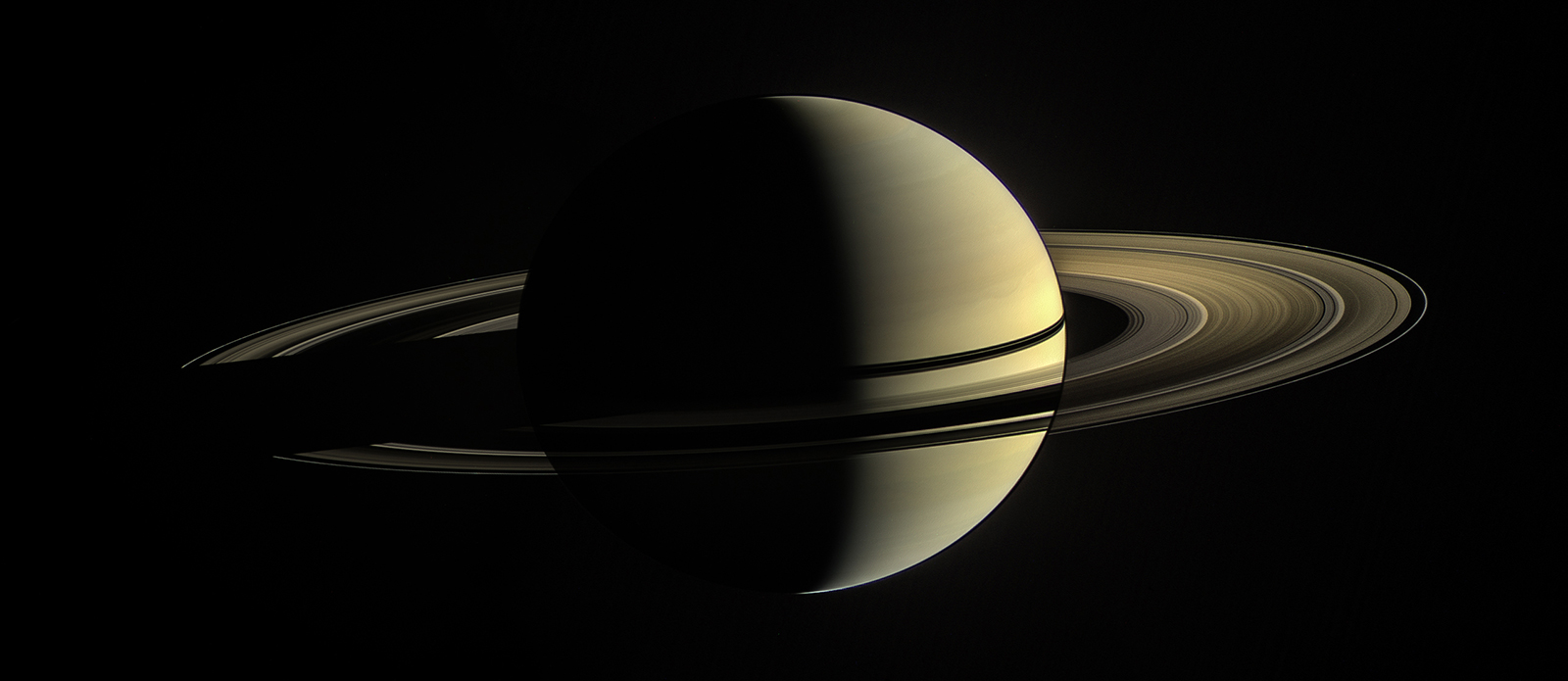 A photograph of Saturn from the Cassini spacecraft, with the left side dark and the right side brightened to reveal ring features. 