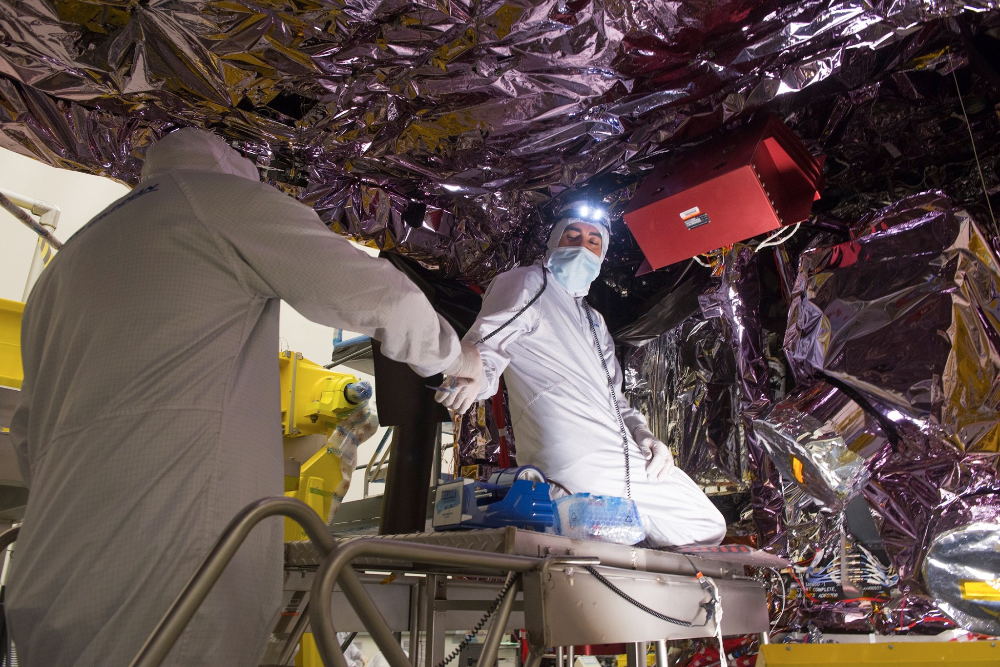 Two people wearing white coverall suits, blue medical masks and white gloves underneath a large piece of space equipment covered in silver thermal blanketing. One person kneels on a raised platform and turns around to accept something from the other person, who stands with their back to the camera. The person facing the camera has a headlamp on their head.