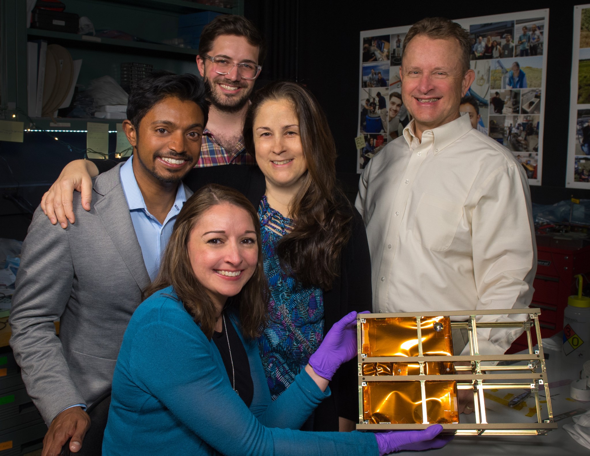 Two women and three men, woman in front is wearing a blue sweater and purple gloves. She is holding technology with a copper color coating on the left and six gold boxes in a 3 x 2 format. 