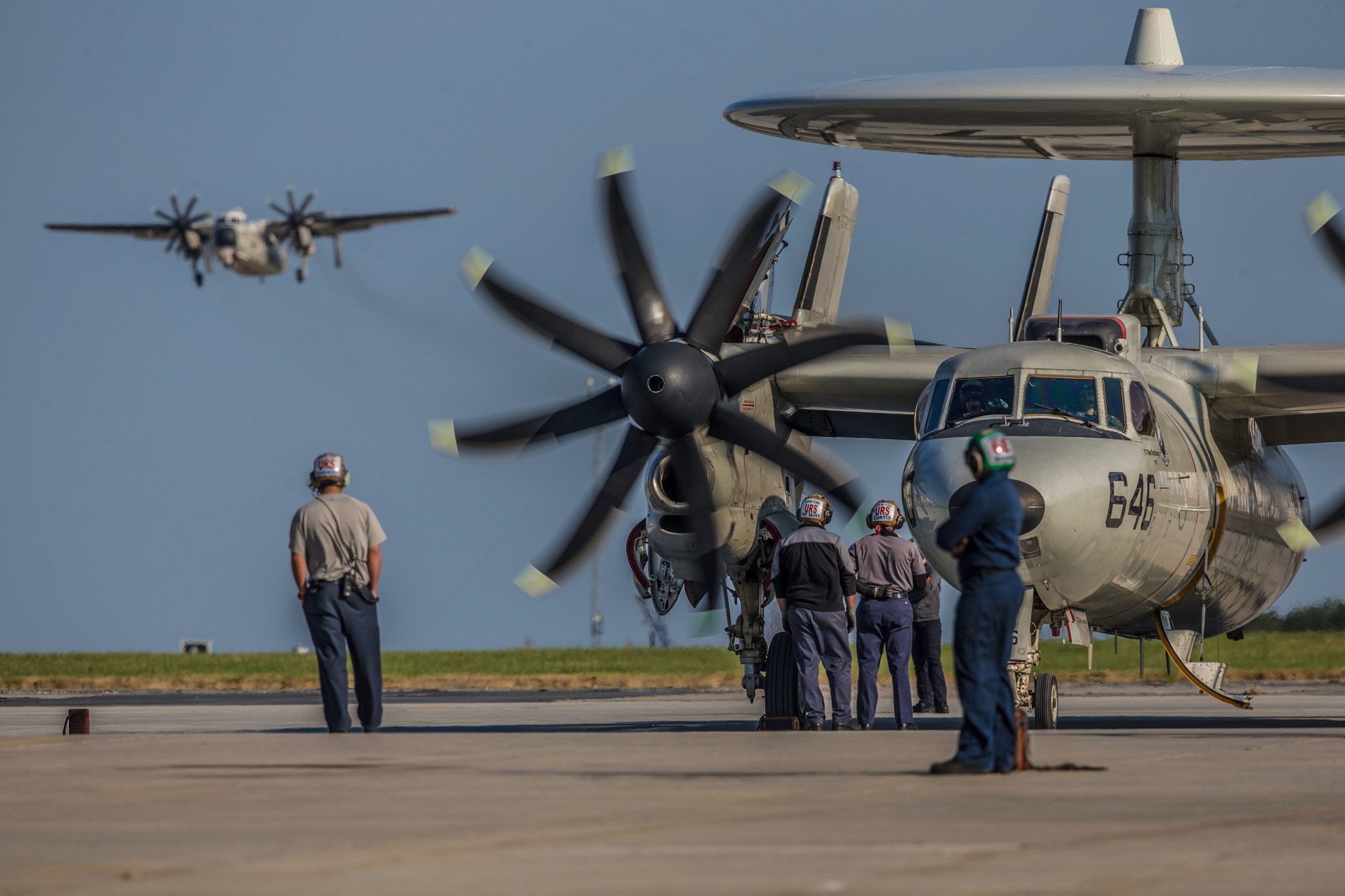 A military aircraft on a tarmac with multiple crew members around. A second military plane is seen in the distance flying in the sky.