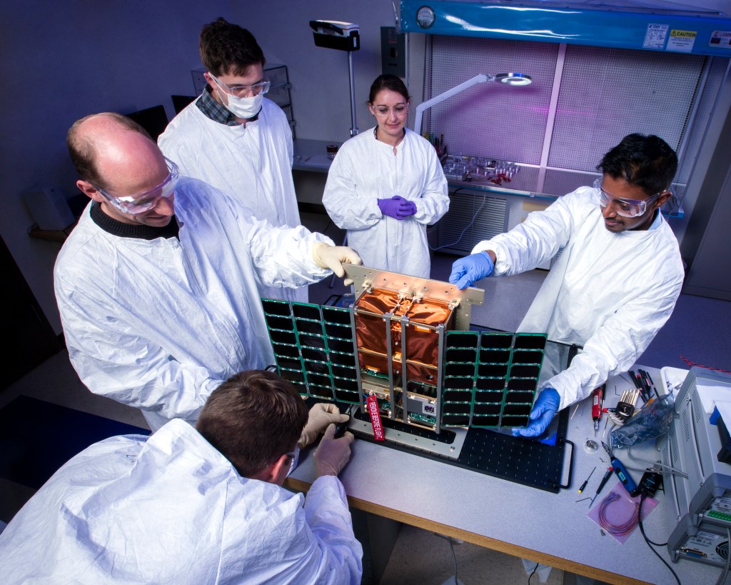 Five people in white lab coats standing around an instrument with a copper colored center and panels on either side.