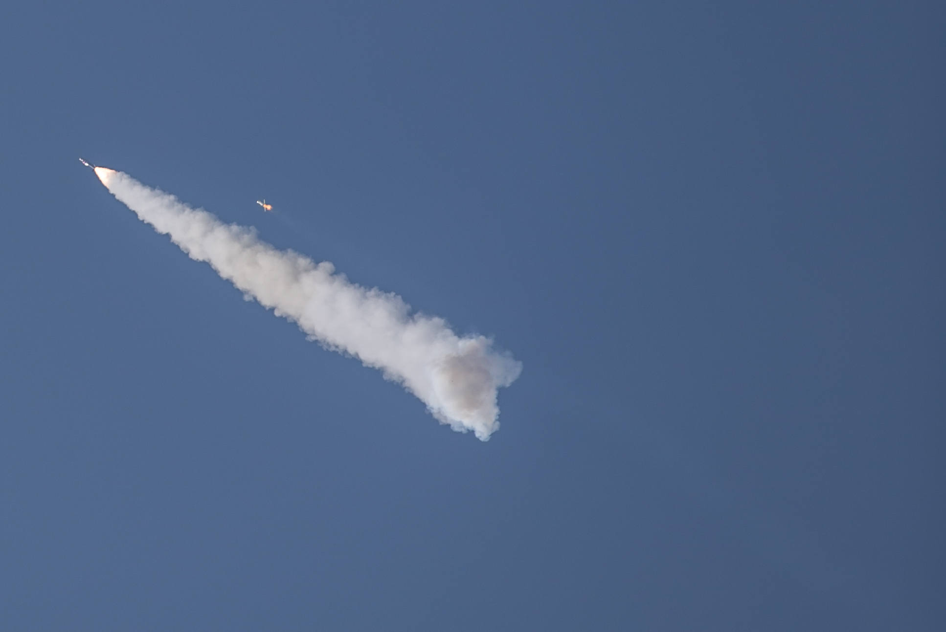 Photo of a sounding rocket stage separation in the sky. The first stage is seen just above the plume cloud, and the second stage with the payload has a plume of smoke under it.