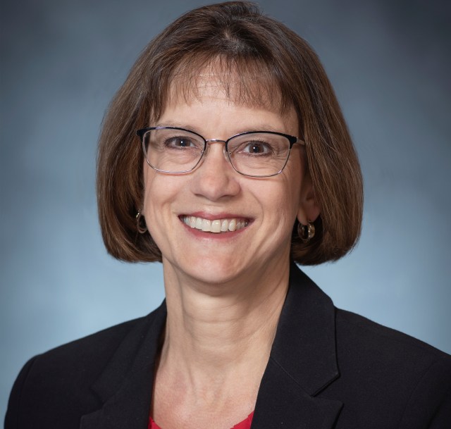 Barbara Esker wearing a dark suit with a red top in front of a gradient gray background.