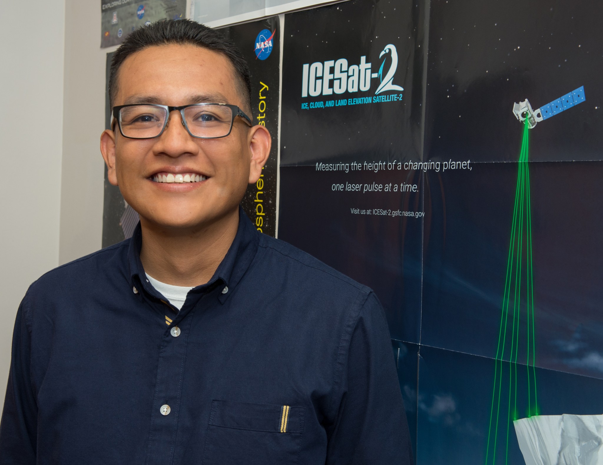 Native American man with black hair and tan skin wears glasses and a navy dress shirt. He stands in front of a poster that says "ICESat 2" with a satellite with green lasers beaming down. 