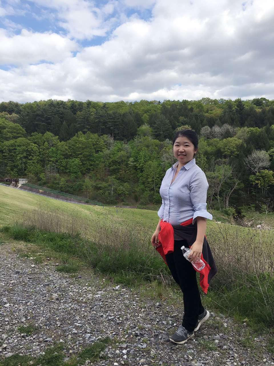 Woman with black hair and light tan skin wears a blue button down, black pants and tennis shoes. She is carrying a water bottle and is hiking on a trail with trees in the background. 