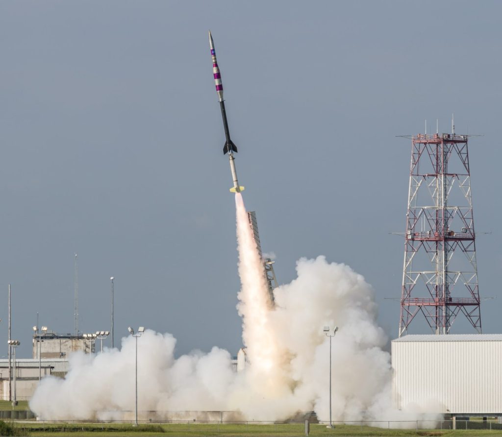 Sounding rocket launching from its pad with a plume of white smoke underneath against a blue sky.