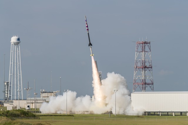 A sounding rocket launches with a bright white plume underneath against a clear, pale blue sky.