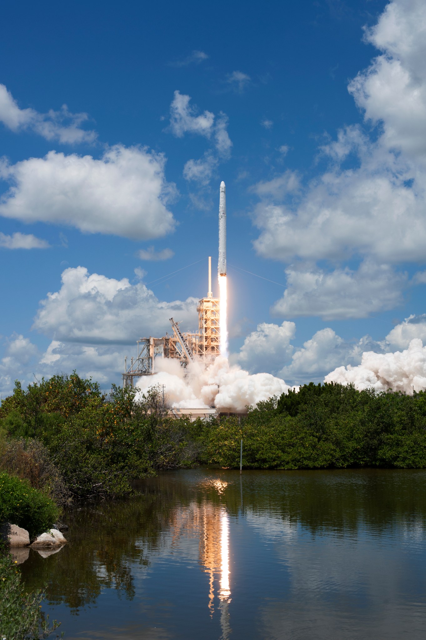  The Falcon 9 launch vehicle lifts off carrying the CRS-12 to the International Space Station. The sky is bright blue with puffy clouds. The rocket shoots up through the center of the frame, with green trees framing its bottom. A body of water in the foreground reflects the rocket, its fire, and the tower.