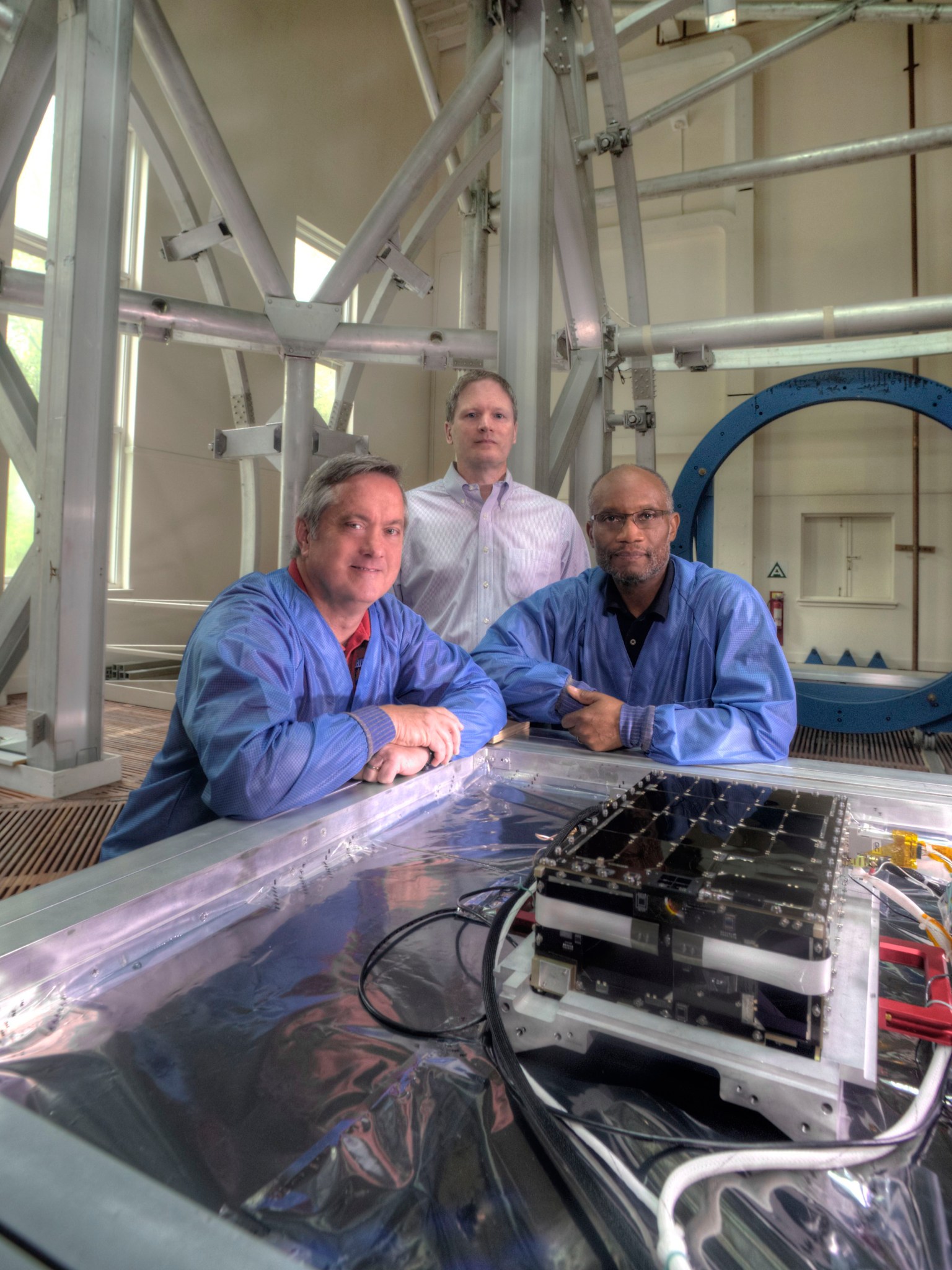 Three men, two in blue lab coats and the third in a dress shirt pose with a black box on a silver lined table with many wires and electrical components. 