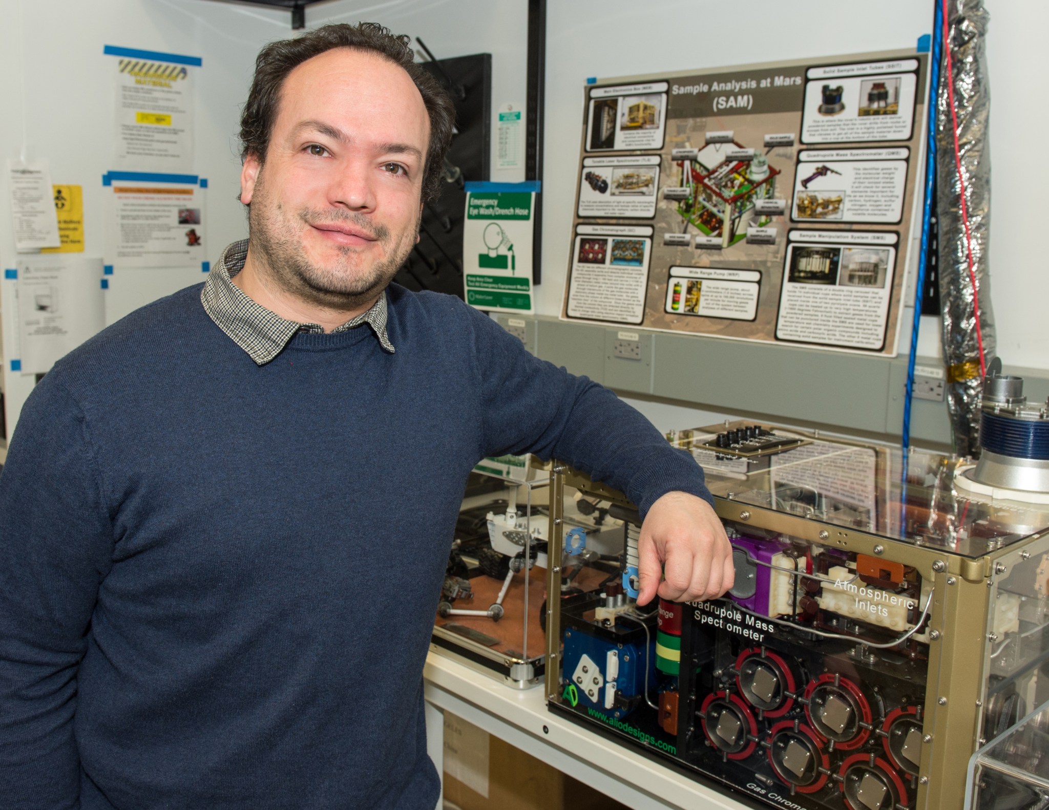 Man with dark brown hair wearing a blue sweater with a collared shirt underneath stands, smiling in front of a Sample Analysis at Mars poster 