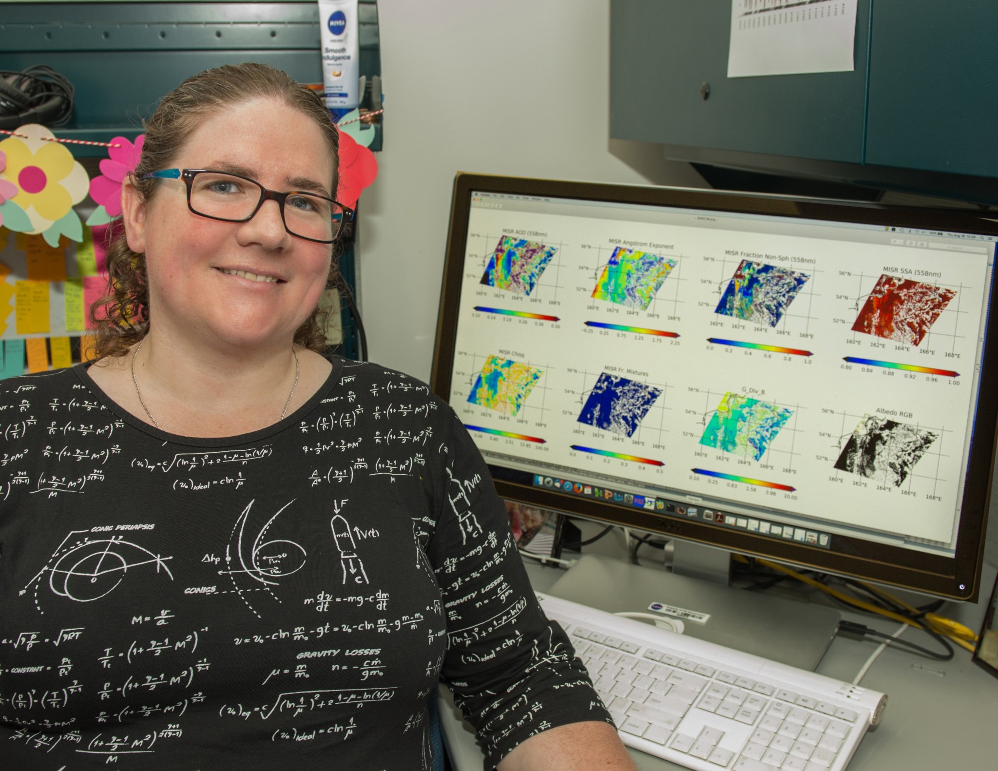 Woman with fair skin and brown curly hair wears her fair in a ponytail. She is wearing a black shirt with math equations and glasses. She is sitting in front of her computer which displays many maps.