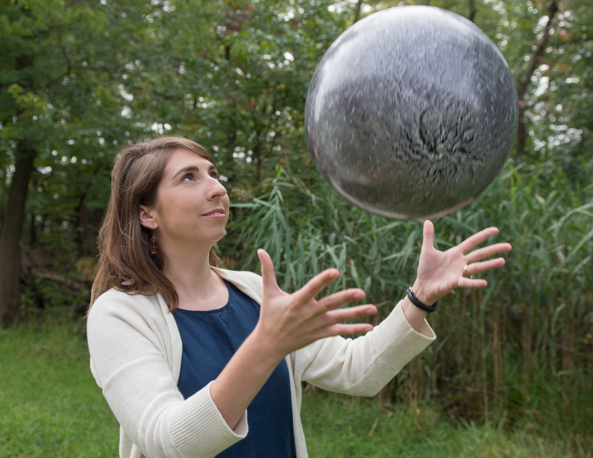 Woman with brown hair and fair skin wears a blue top and cream sweater. She is outside in front of trees and is tossing a model of the Moon into the air. 