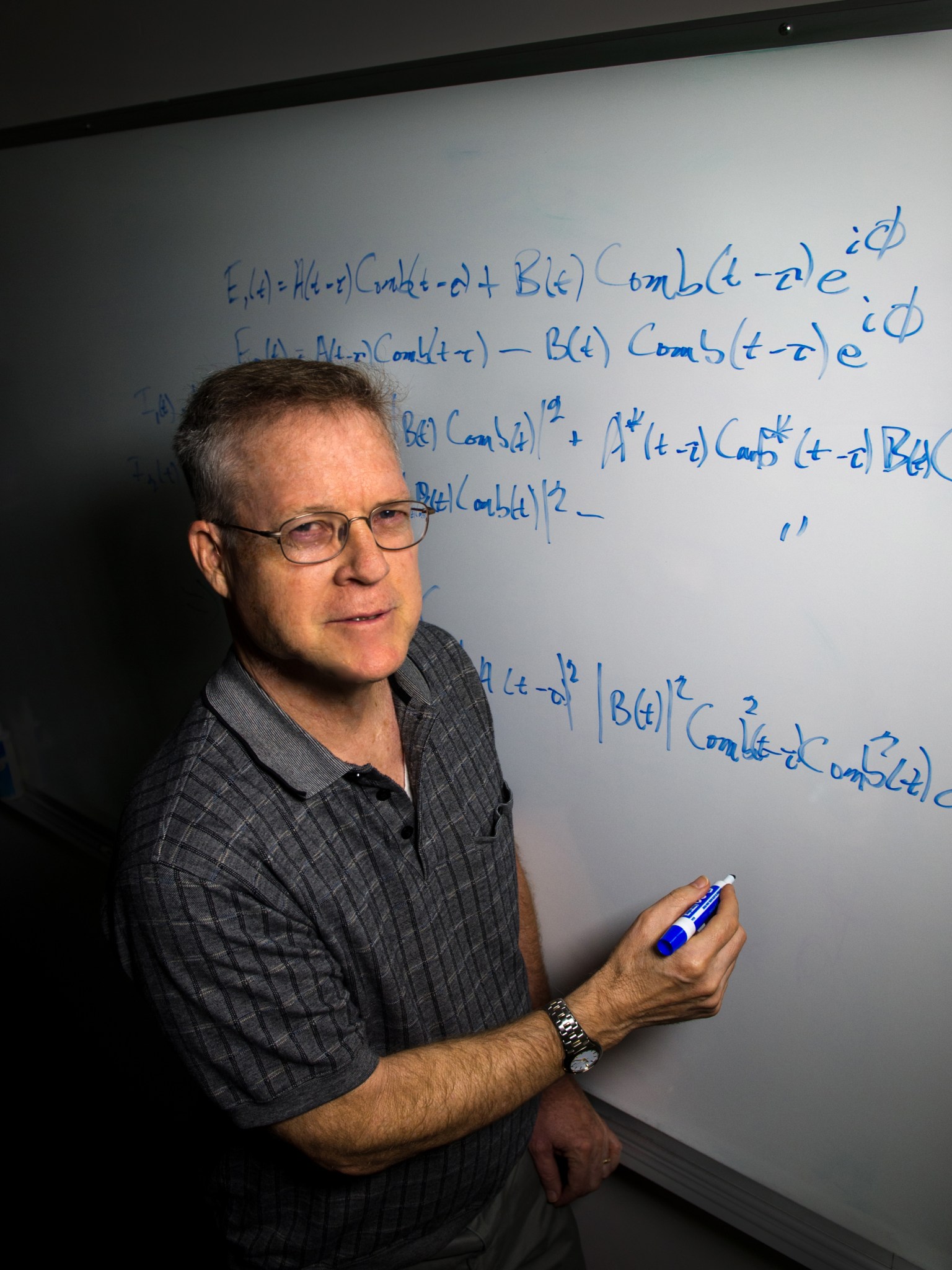 Man with fair skin and short grey-brown hair wears glasses and a grey polo. He is holding a blue dry erase marker and standing next to a white board with many equations written. 