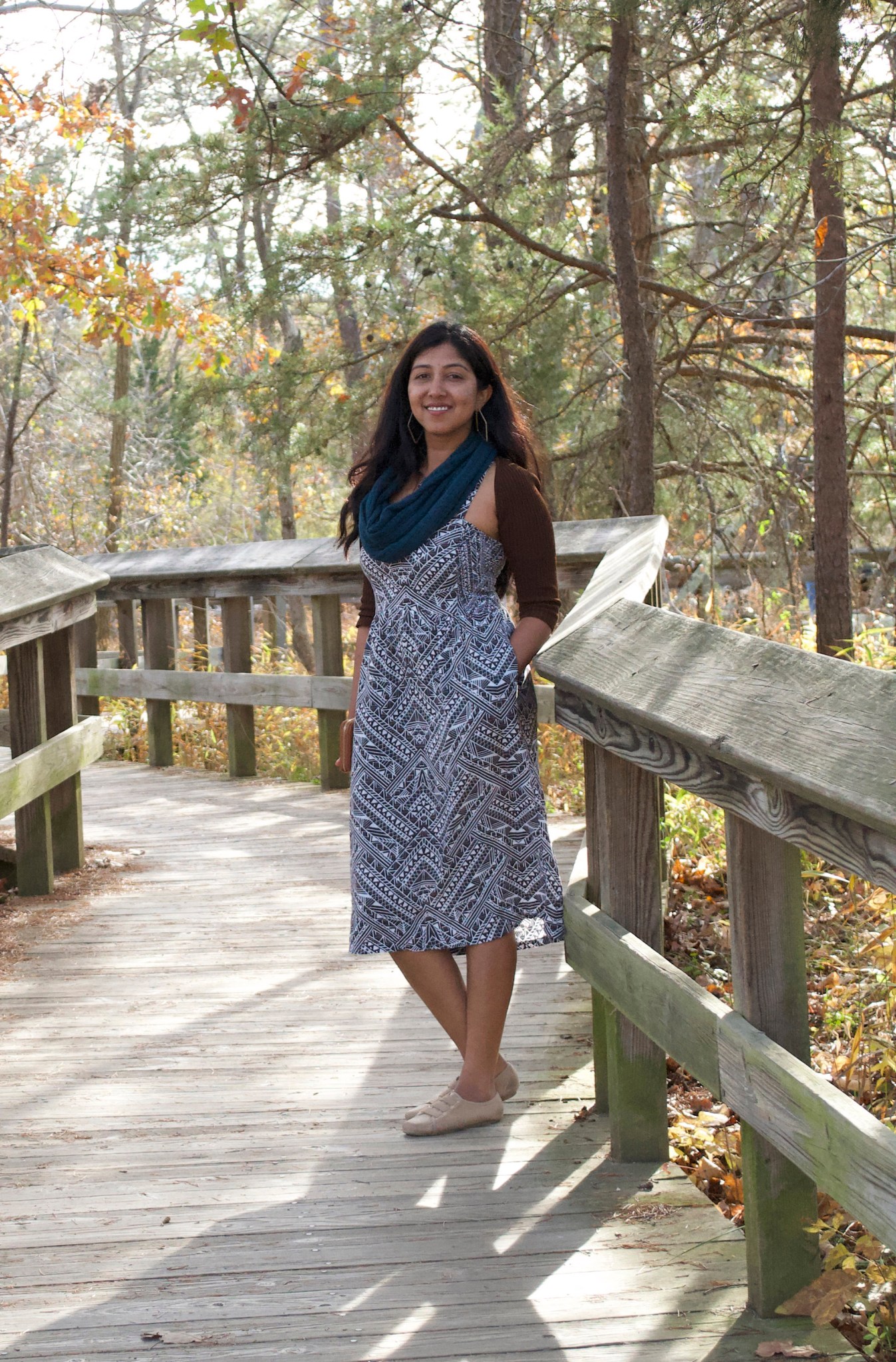 Woman with tan skin and long dark brown hair wears a black and white dress with a brown sweater and a teal scarf. She is smiling and standing on a wooden walkway with trees surrounding her. 
