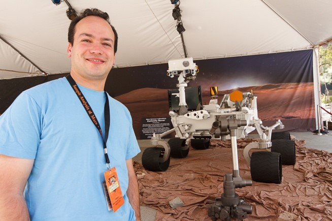 Man with dark brown hair wears a light blue shirt and an orange badge. He stands in front of a model of the Mar's Curiosity rover.