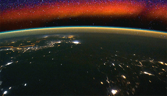 The dark limb of Earth sliding under the camera on the Space Station. Electric lights glow on land. Above the limb of Earth, the atmosphere glows a gold and blue, forming a bubble above Earth. Above that, airglow shines as reddish orange and purple lights.