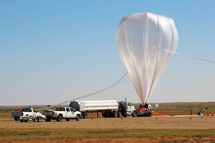 A large scientific balloon is seen inflated, tethered and sitting next to several vehcles in a field.