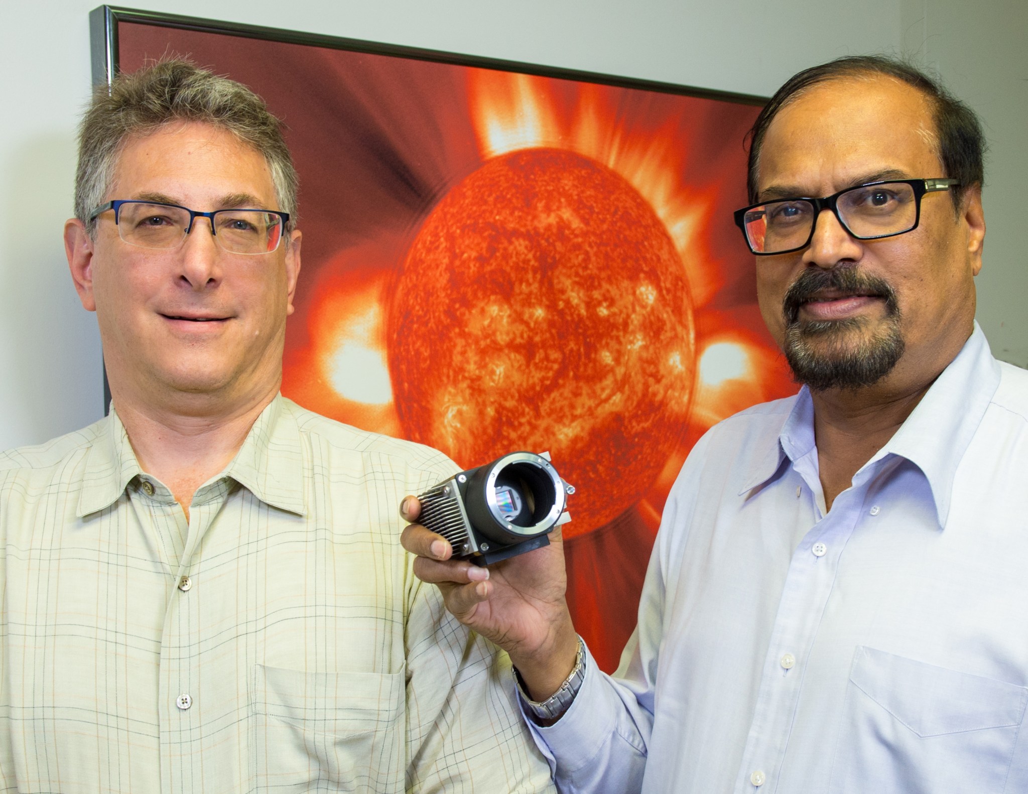 Heliophysicists Jeff Newmark (left) and Nat Gopalswamy (right) standing in front of a poster of a Sun image and holding a polarization camera