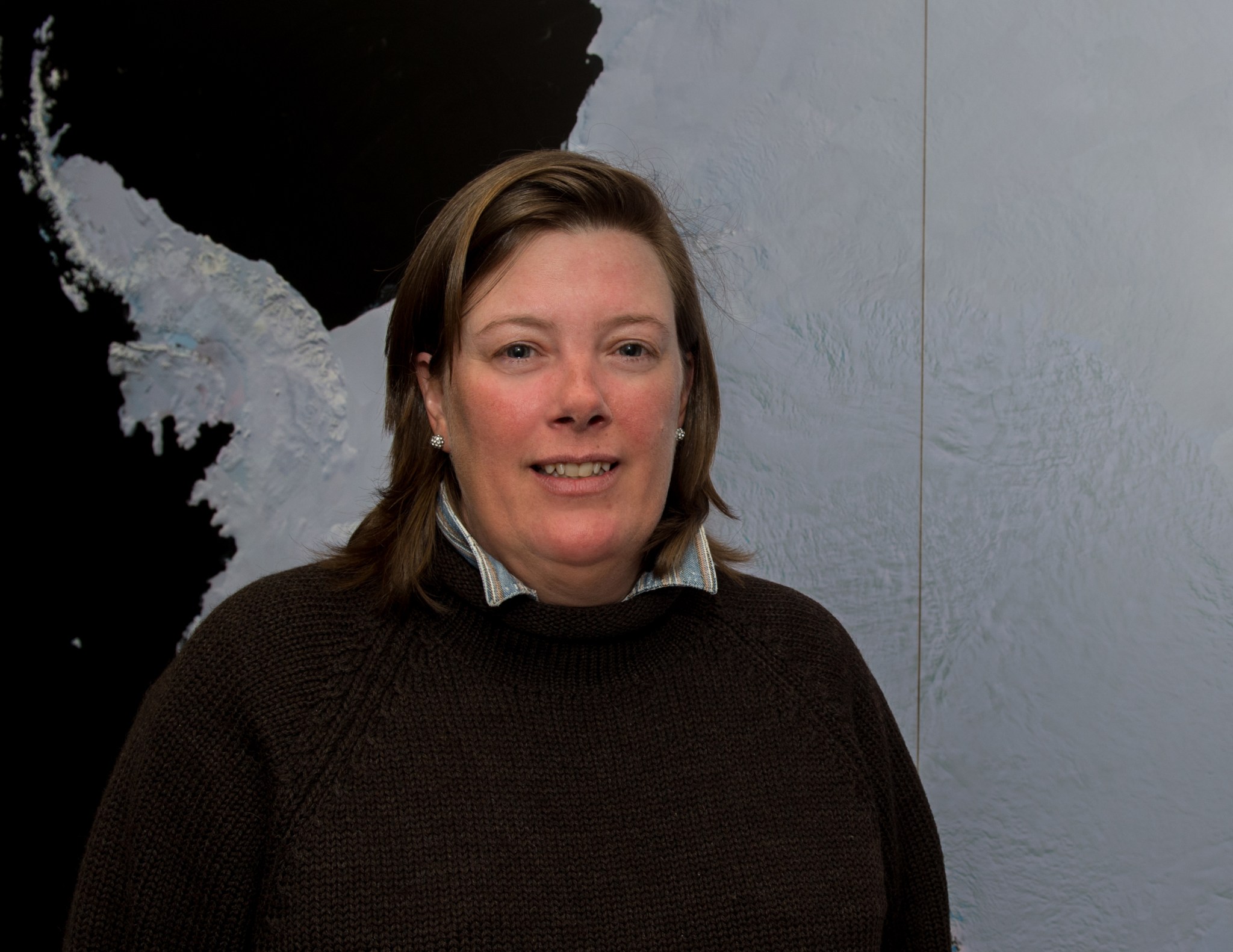 Woman with short brown hair wears a brown sweater with a collared shirt underneath. She stands in front of a map of ice.