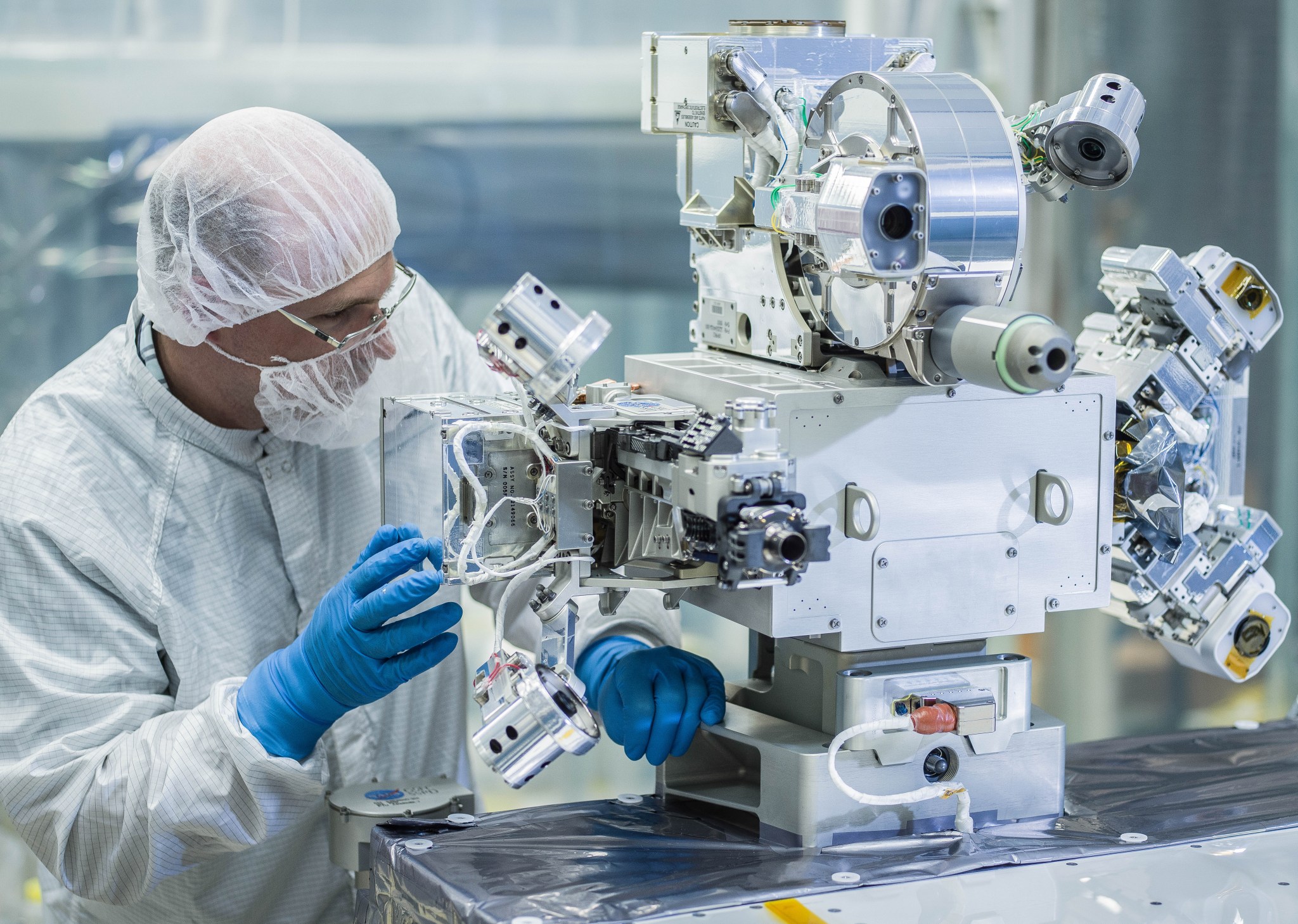 Engineer in white clean room suit with blue gloves performs fit check on RRM3 tools. He is looking at a silver instrument with several protruding features settled on a table.