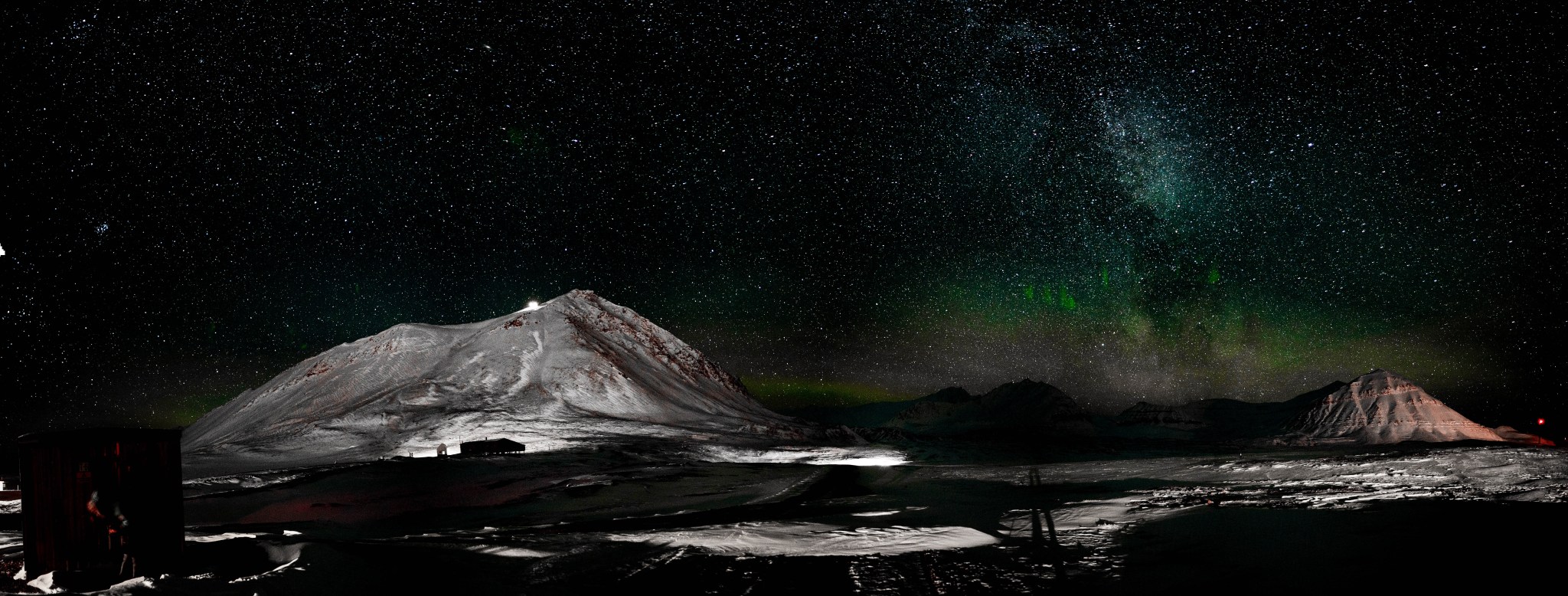 A panoramic photo of the sky at night, appearing mostly black and white except for a blush of green and blue near the horizon indicating the aurora borealis. Whitish peaks of snow jut up like small mountains near the left and right of the image, and the shadow of a person is visible near the center.