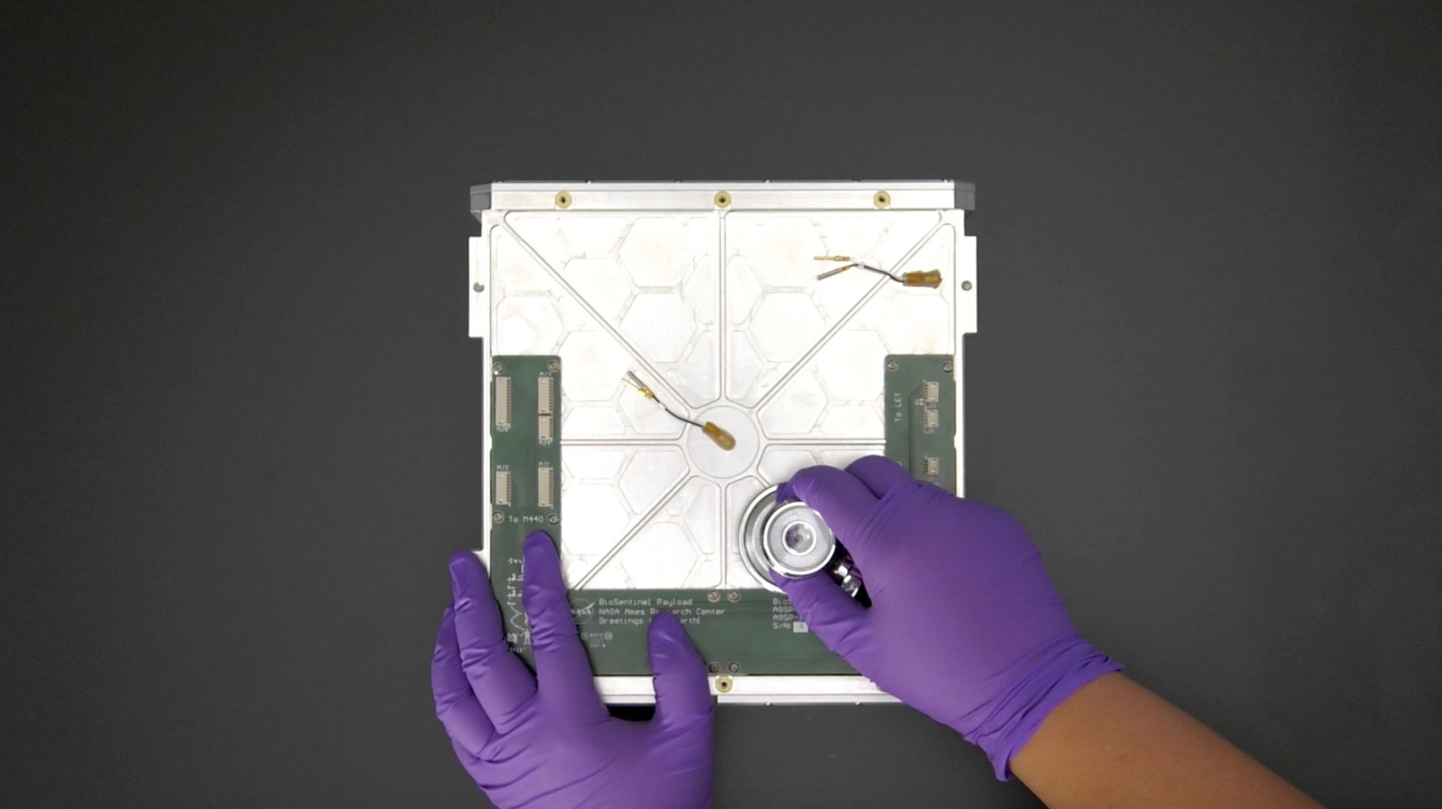 Hands wearing purple gloves are seen holding a stethoscope up to a piece of hardware from a satellite.
