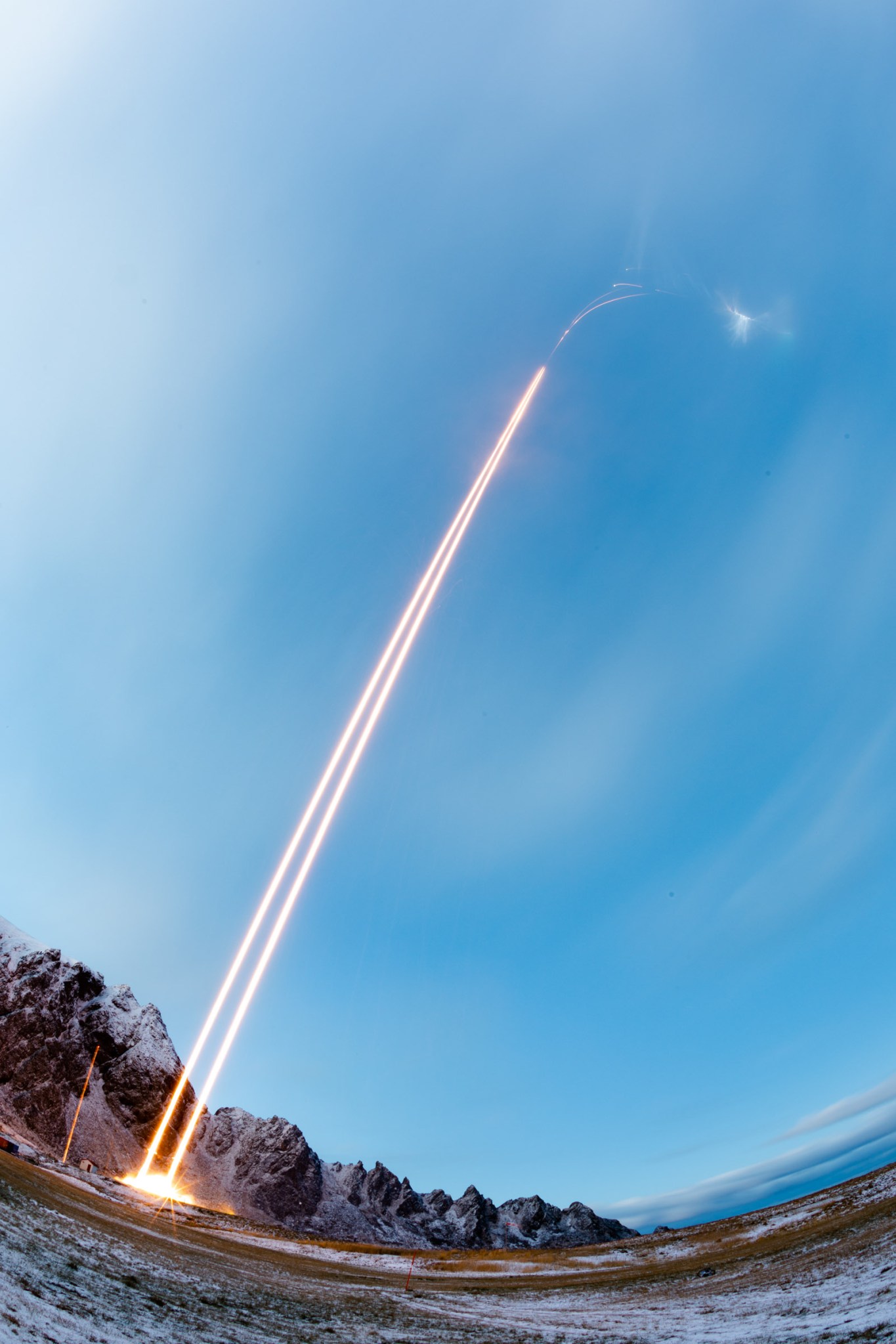 A long-exposure image of two sounding rockets represented by two bright yellow lines launching into a blue sky.
