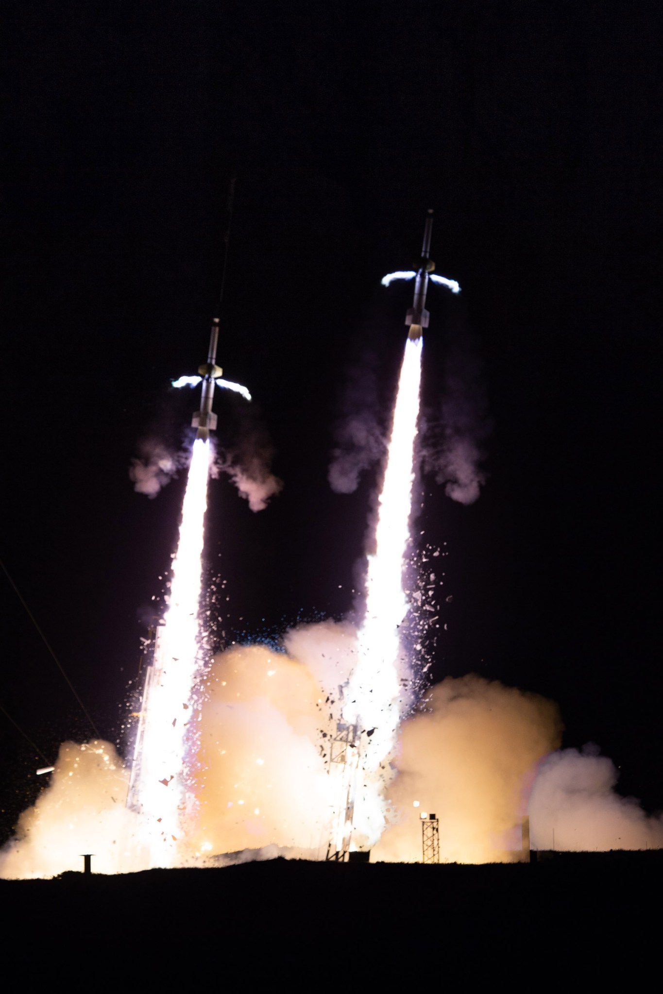 A composite image of two rockets that launched separately put together into one image. The background is black and the two rockets are each in mid-launch, with a white plume of smoke underneath each. Each have a little flame on either side in the middle of the rocket, representing the spin motors firing.