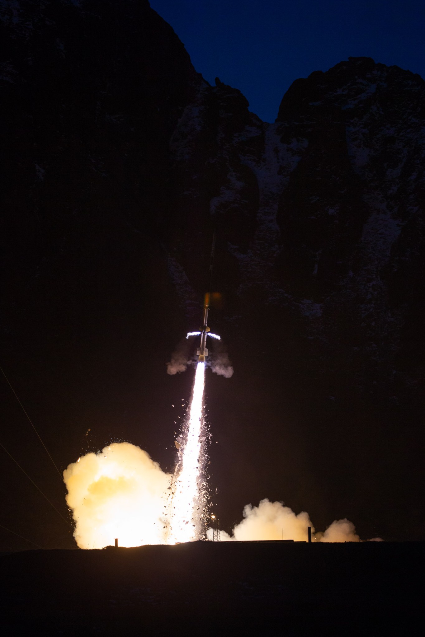 A rocket blasts off into the night, with a bright tail of exhaust and billowing plumes below it.