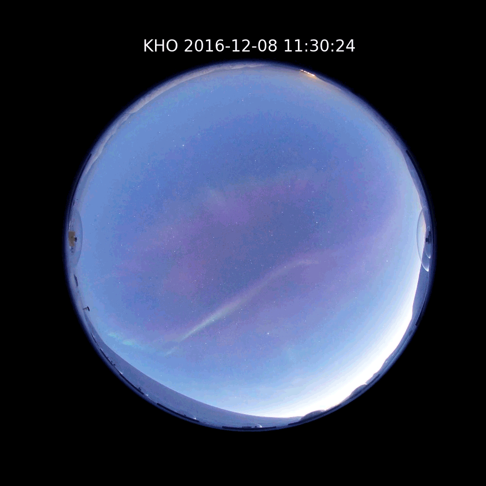 A round, all-sky camera view shows the daytime sky in blue with purple and green streaks of aurora waving and shimmering across the sky.