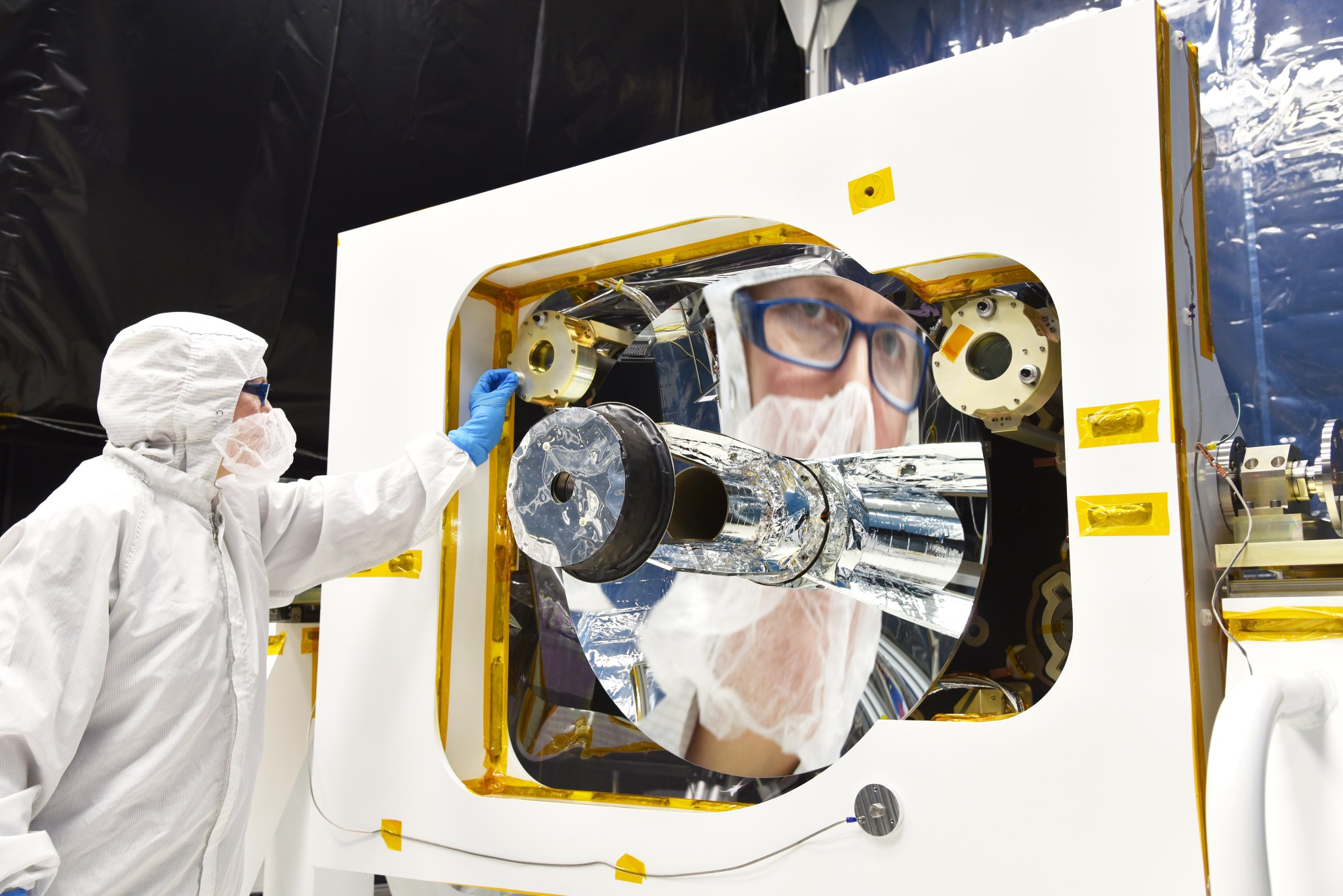 GEDI receiver telescope in the clean room, scientist wearing clean room suit looking into mirror and reflection is shown much larger. The scientist is wearing blue glasses and the mirror is round with an instrument in the center. 