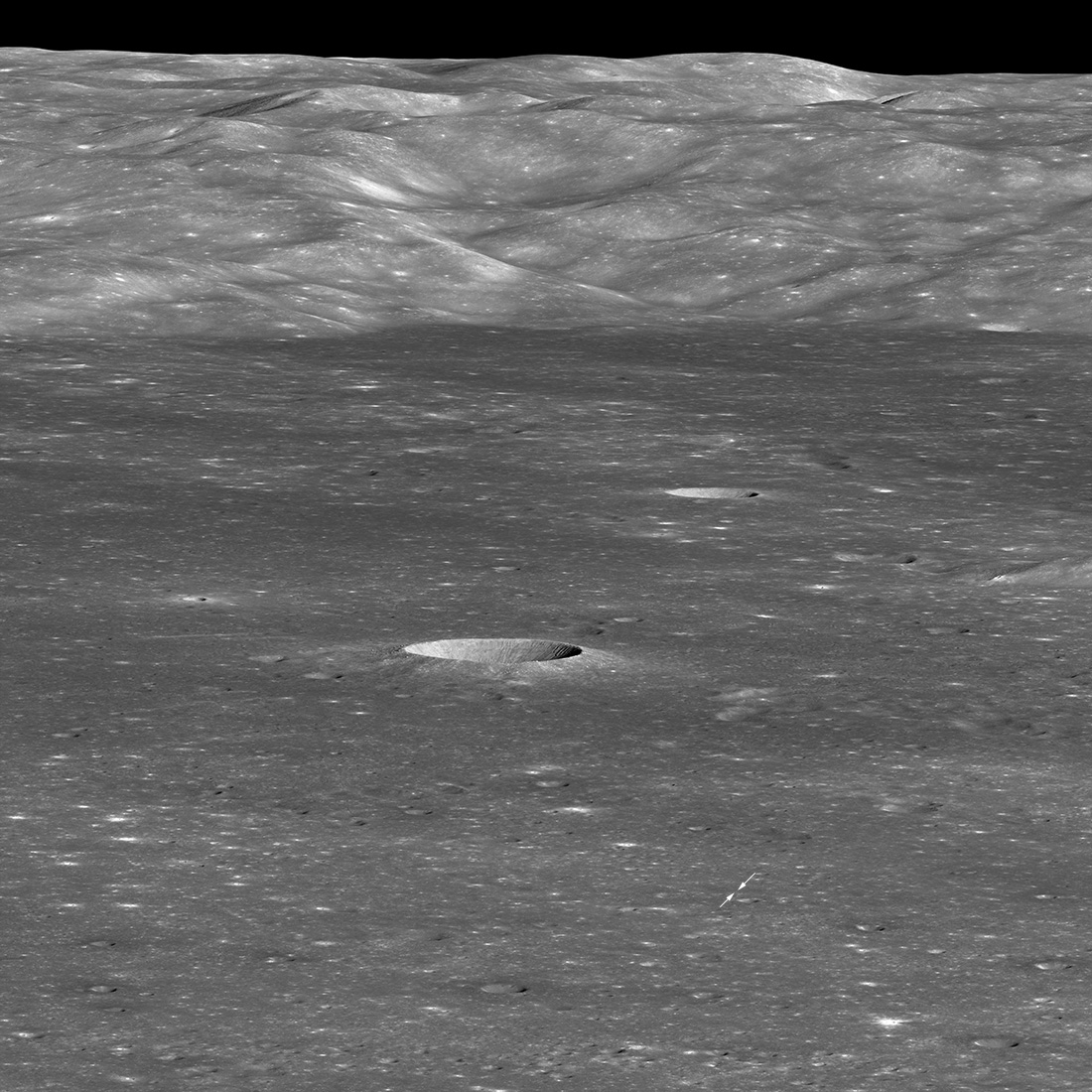 A grayscale photo of the surface of the Moon, showing the landing site of the Chinese spacecraft Chang'e 4. A large crater is prominent in the center of the image and a mountain range leading to a black sky at the top. The lunar surface is mostly flat, with many small craters and some larger ones visible. leading up to the mountains, which gradually change from foothills to tall mountains near the top edge. Below and to the right of the center crater, two tiny white arrows show the Chang'e landing site.