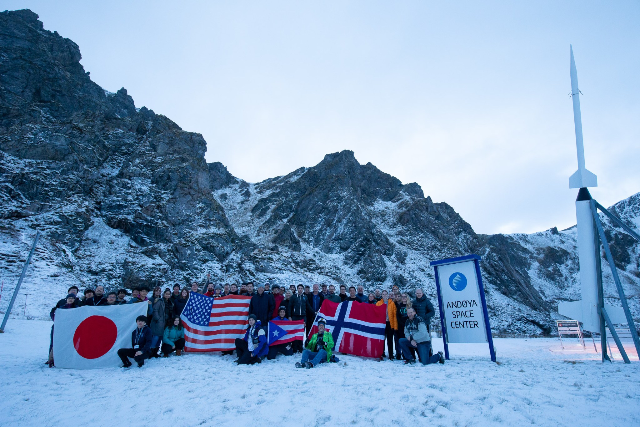 G-CHASER student teams at the Andøya Space Center. Several people bundled in winter clothes stand and sit in a group in the snow. Behind them, mountains have patches of snow. The people are in a snowy field holding various flags.