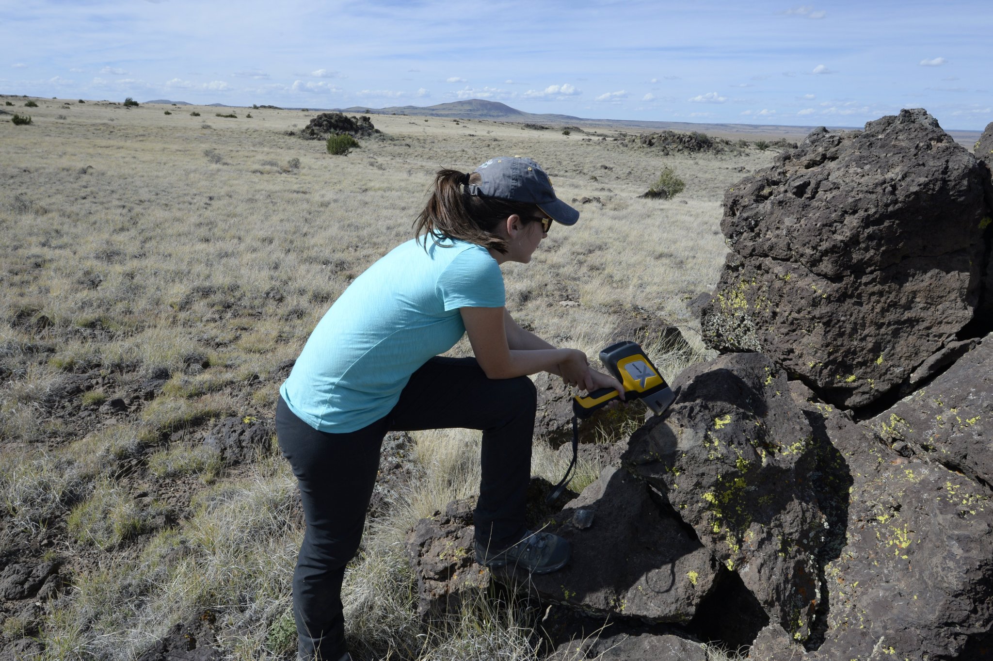 Woman wearing a blue shirt and a hat holds a spectrometer and squats beside black volcanic rock. 