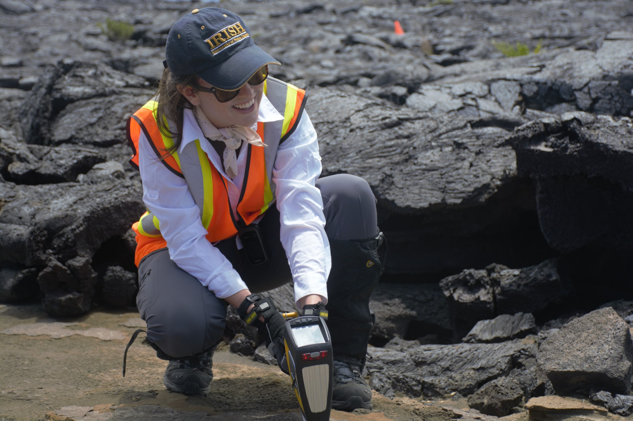 Woman wearing a white button down, orange safety vest and a hat holds a spectrometer and squats beside black volcanic rock. 