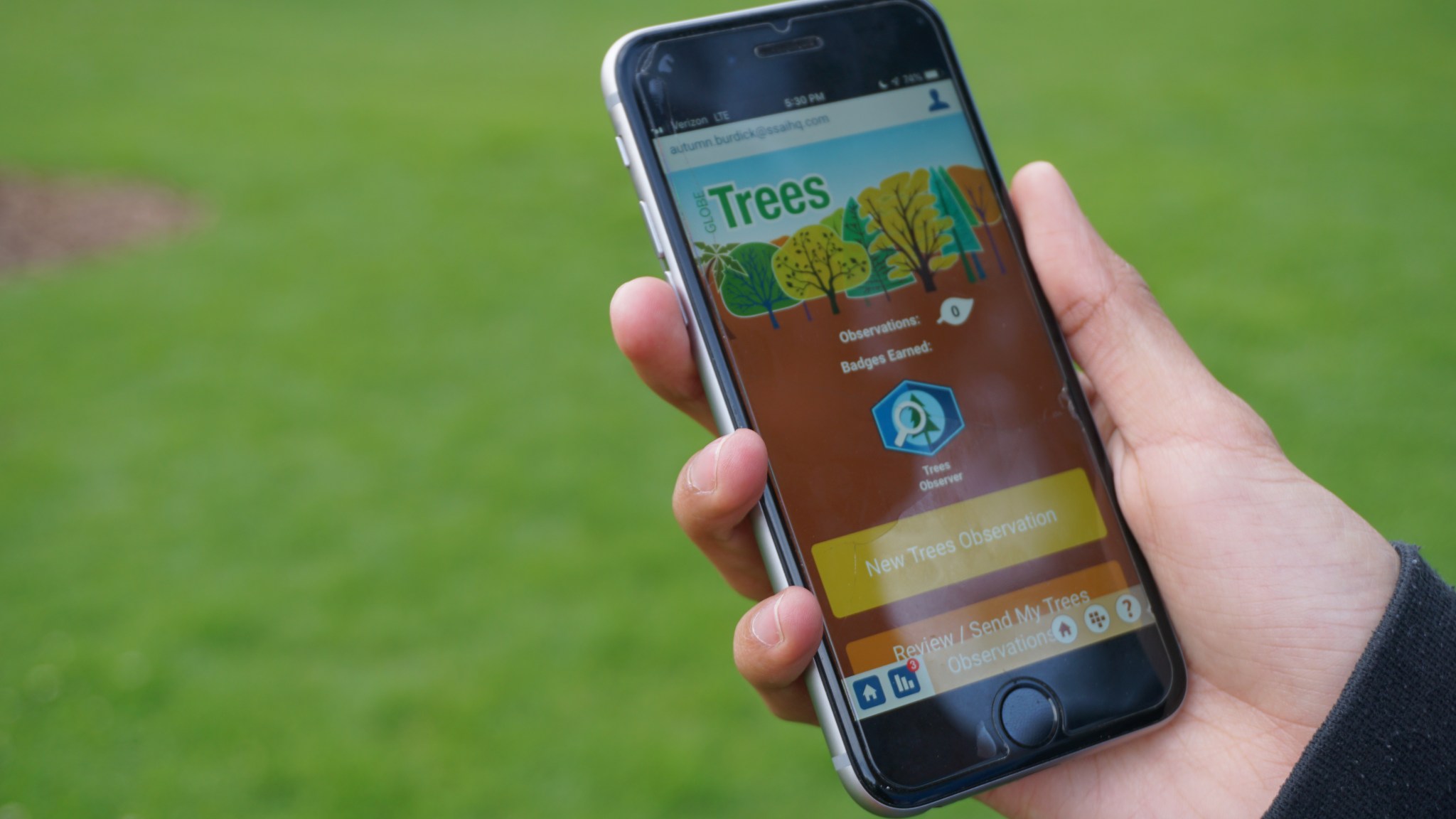 A hand holding an iPhone. The screen shows the GLOBE Trees app, with a button that says "New Trees Observation."