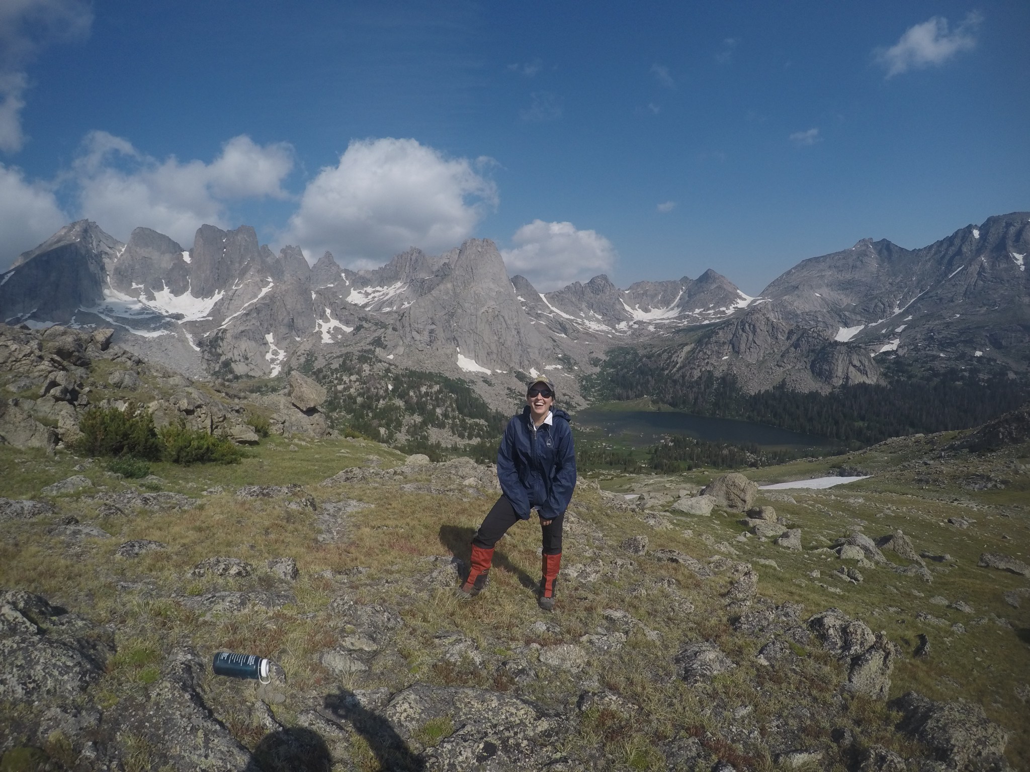 Woman wears a blue rain jacket and red boots. She is smiling in front of a beautiful mountain landscape. 