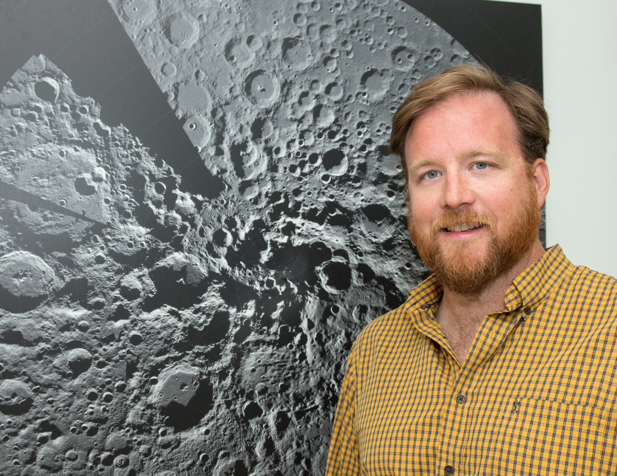 Man with reddish brown hair and facial hair stands next to an image of the Moon's surface wearing a yellow button down shirt.