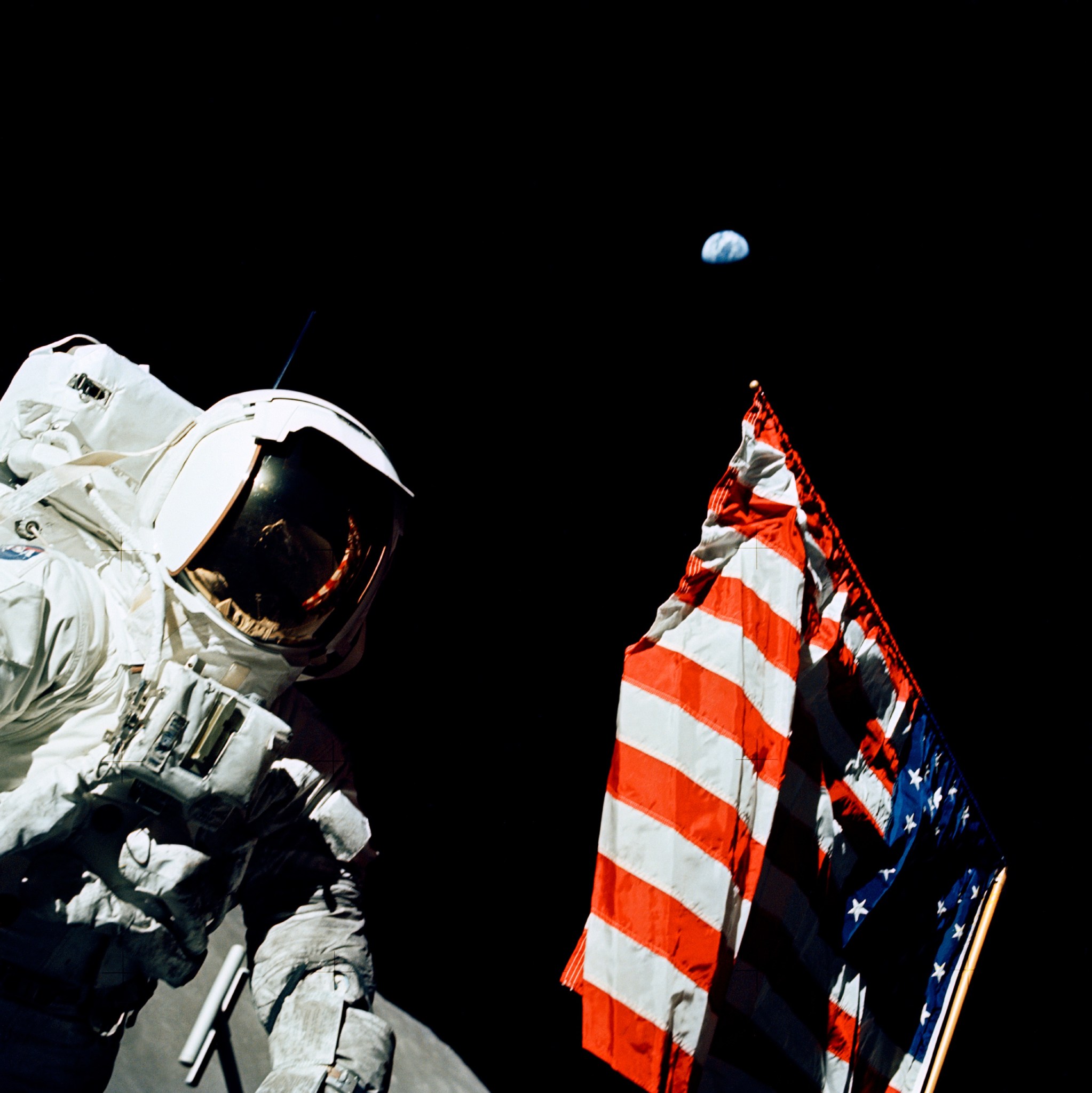 Geologist-Astronaut Harrison Schmitt, Apollo 17, is photographed next to the American Flag during final Moon shot. Earth is visible as a tiny blue dot in the distance. 
