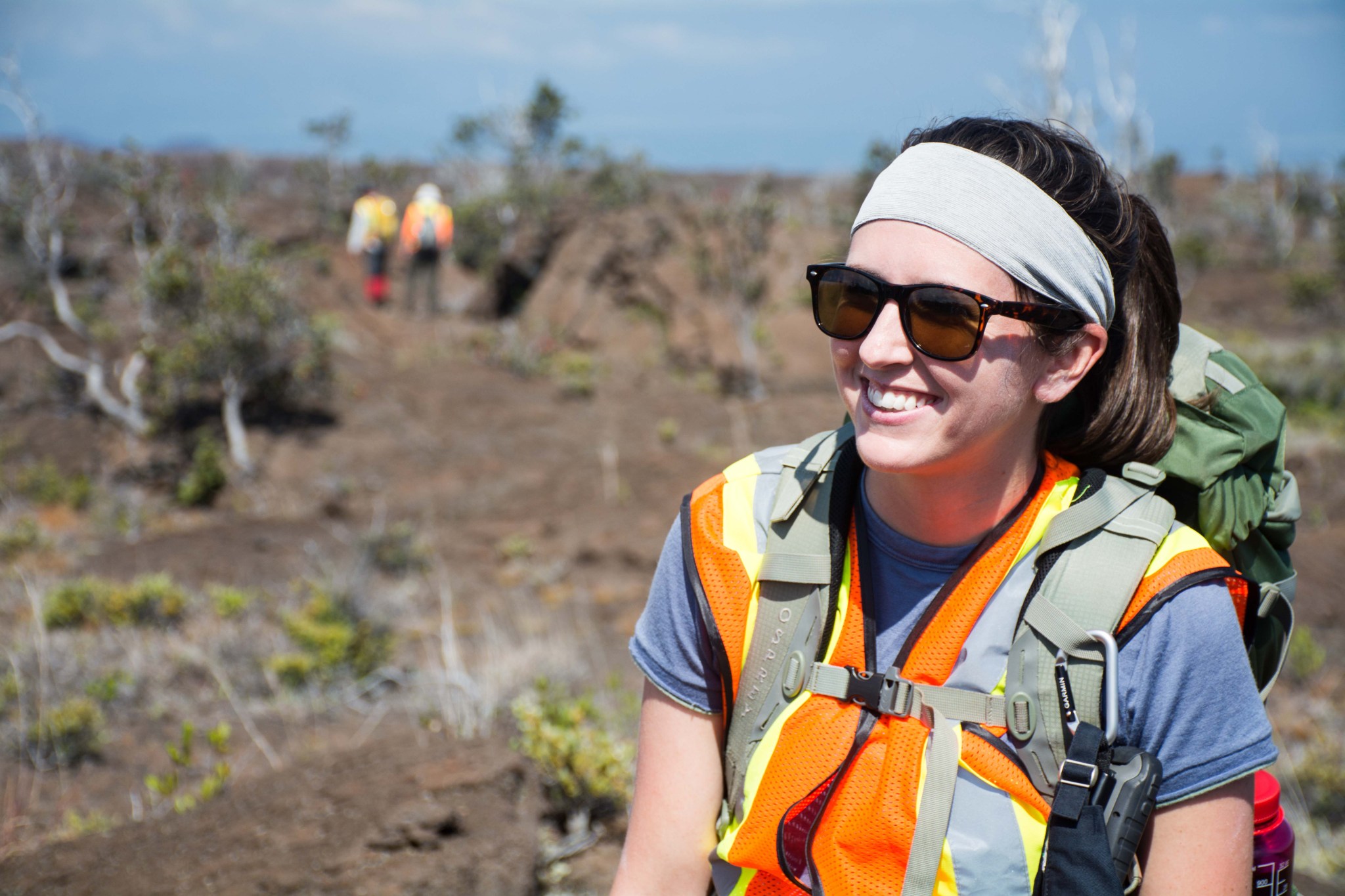 Woman with brown hair in a ponytail wears sun glasses and a grey headband. She is also wearing a blue t shirt, orange safety vest and a green backpack. She's standing outside surrounded by small trees. 
