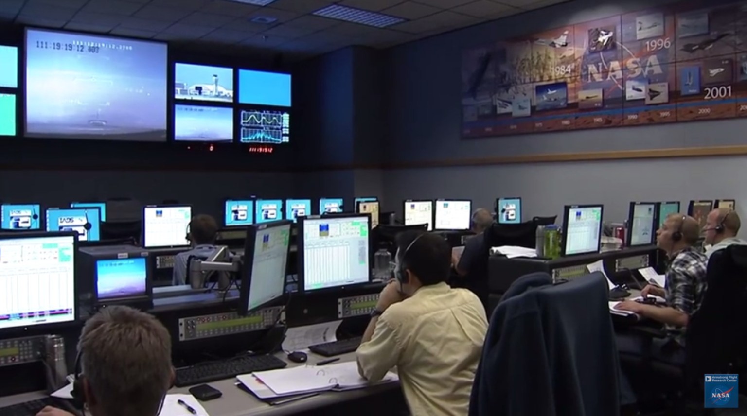 Mission control room with rows of computer stations. Six people sit at six of the many stations. A large monitor on the wall at the front of the room shows nine video feeds, four small video feeds on either side of one large video feed in the center.