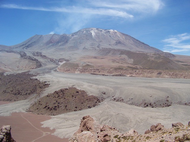 Image of Lascar Volcano in Chile. The volcano is brown with white on the top and the sky is blue with wispy, white clouds