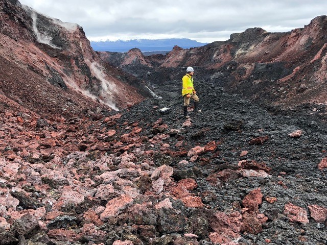 Man wears a hard hat, yellow jacket, and boots and stands in a lava channel of ash and red rocks. 